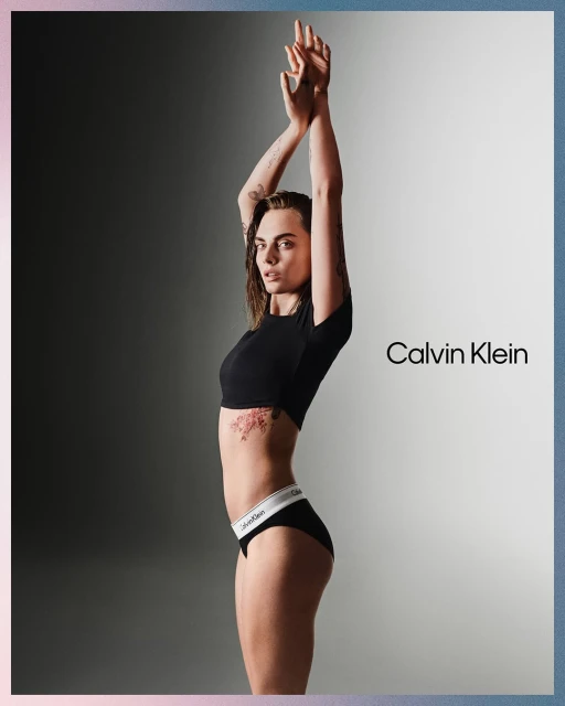 A slender, tanned adult female with a tattoo on her torso poses in black underwear and a crop top in a grey gradient studio shot.
