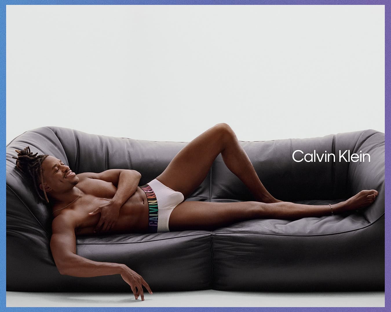A shirtless African-American man with dreadlocks wearing Calvin Klein underwear, laying on a grey leather couch against a white backdrop.