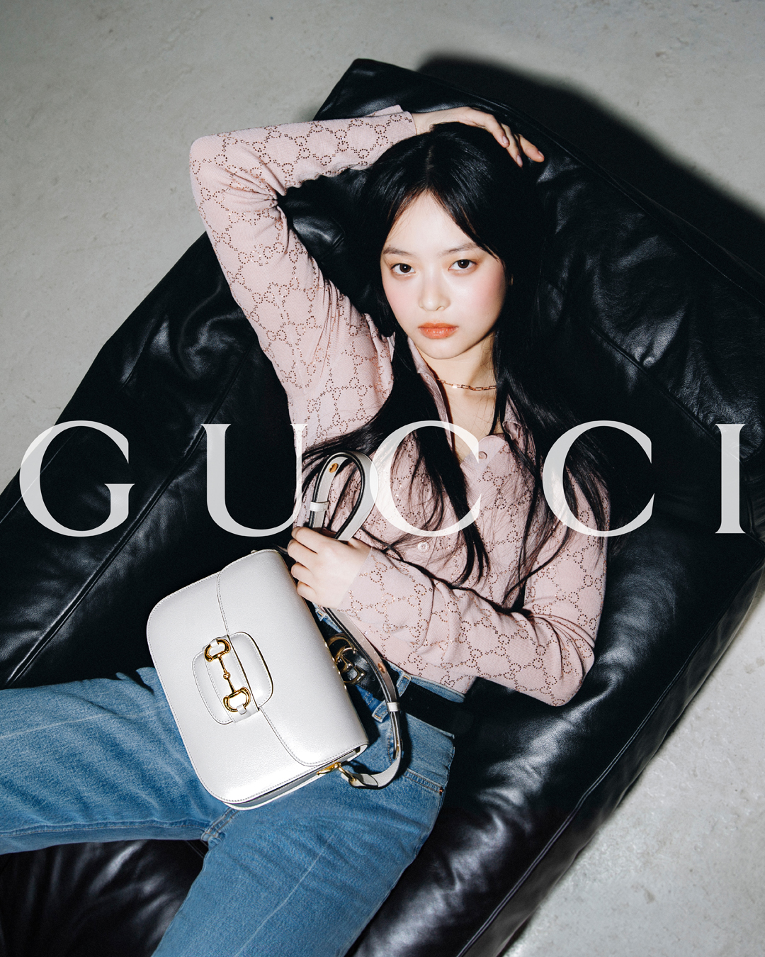A brunette woman poses with a white Gucci purse on a black leather couch, wearing a pink cardigan and blue jeans in an indoor studio.