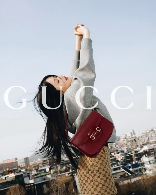 Fashion shot of a woman in a gray sweatshirt and skirt with a red purse, posing with her arms up in an urban setting.