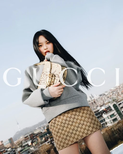 A woman poses with a Gucci purse in an urban outdoor scene with a light sky, wearing a gray sweatshirt and patterned skirt with the Gucci logo.