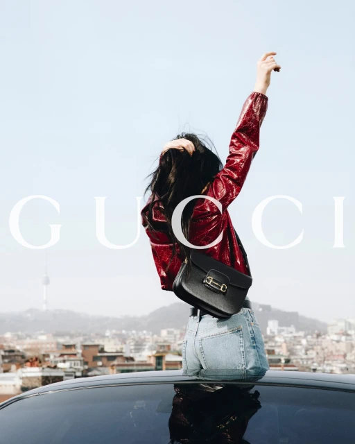 Woman in red jacket and blue jeans stands on car in city, holding her arm up, with Gucci logo visible in the composition.
