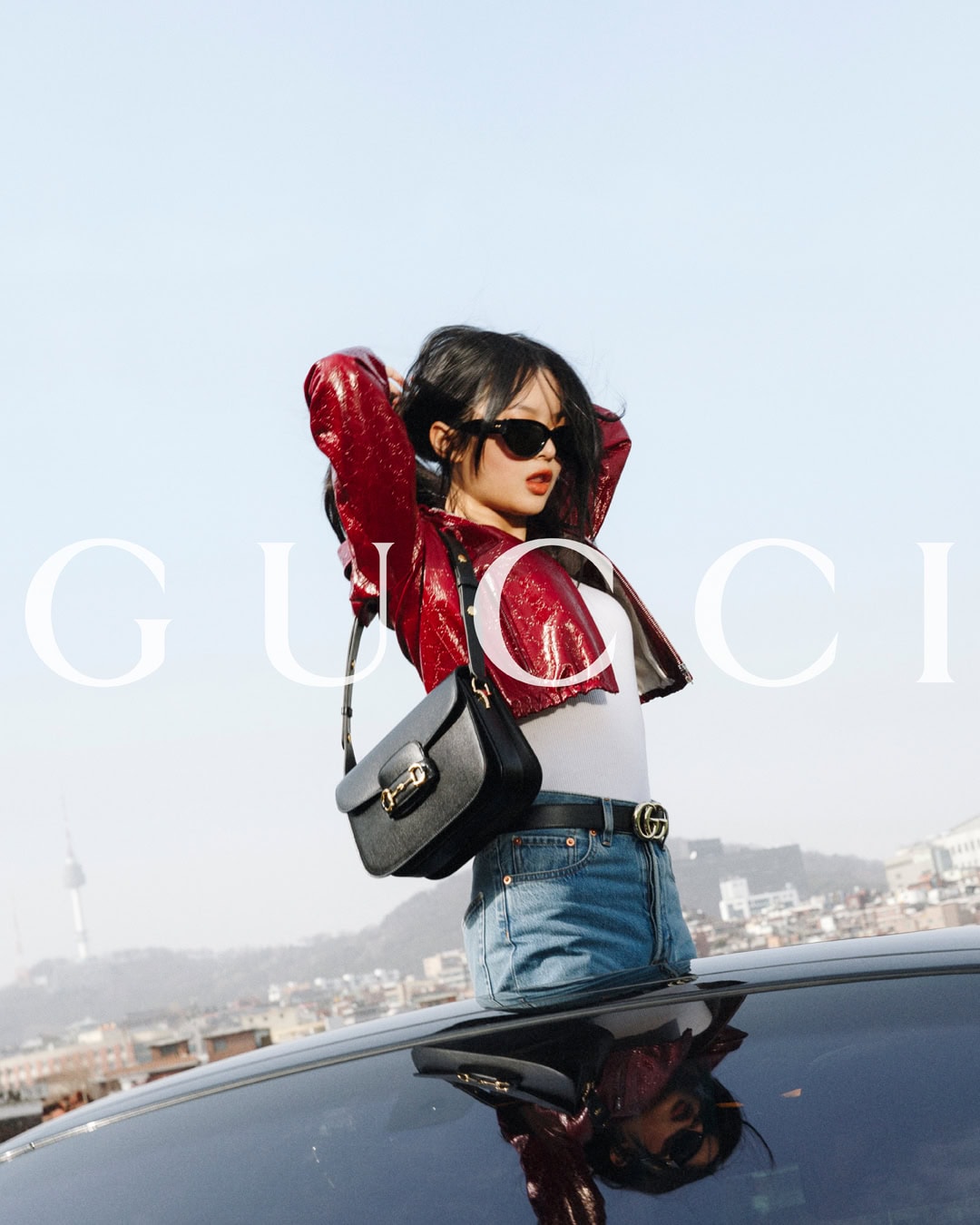 A woman wearing a red jacket and jeans poses on a car roof in an urban setting with the Seoul Tower visible in the background.