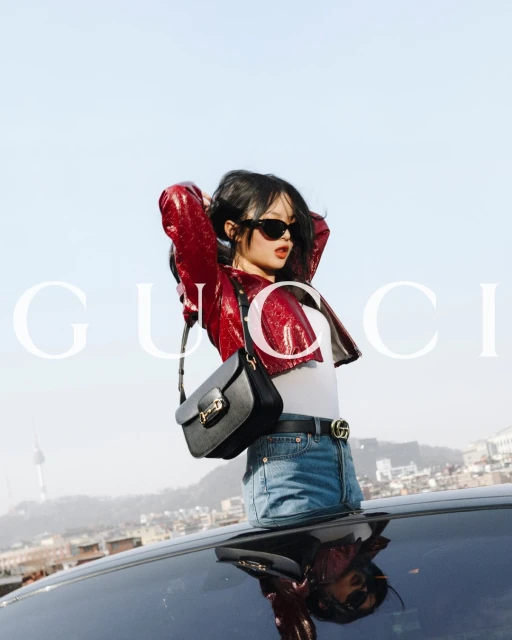 A woman wearing a red jacket and jeans poses on a car roof in an urban setting with the Seoul Tower visible in the background.