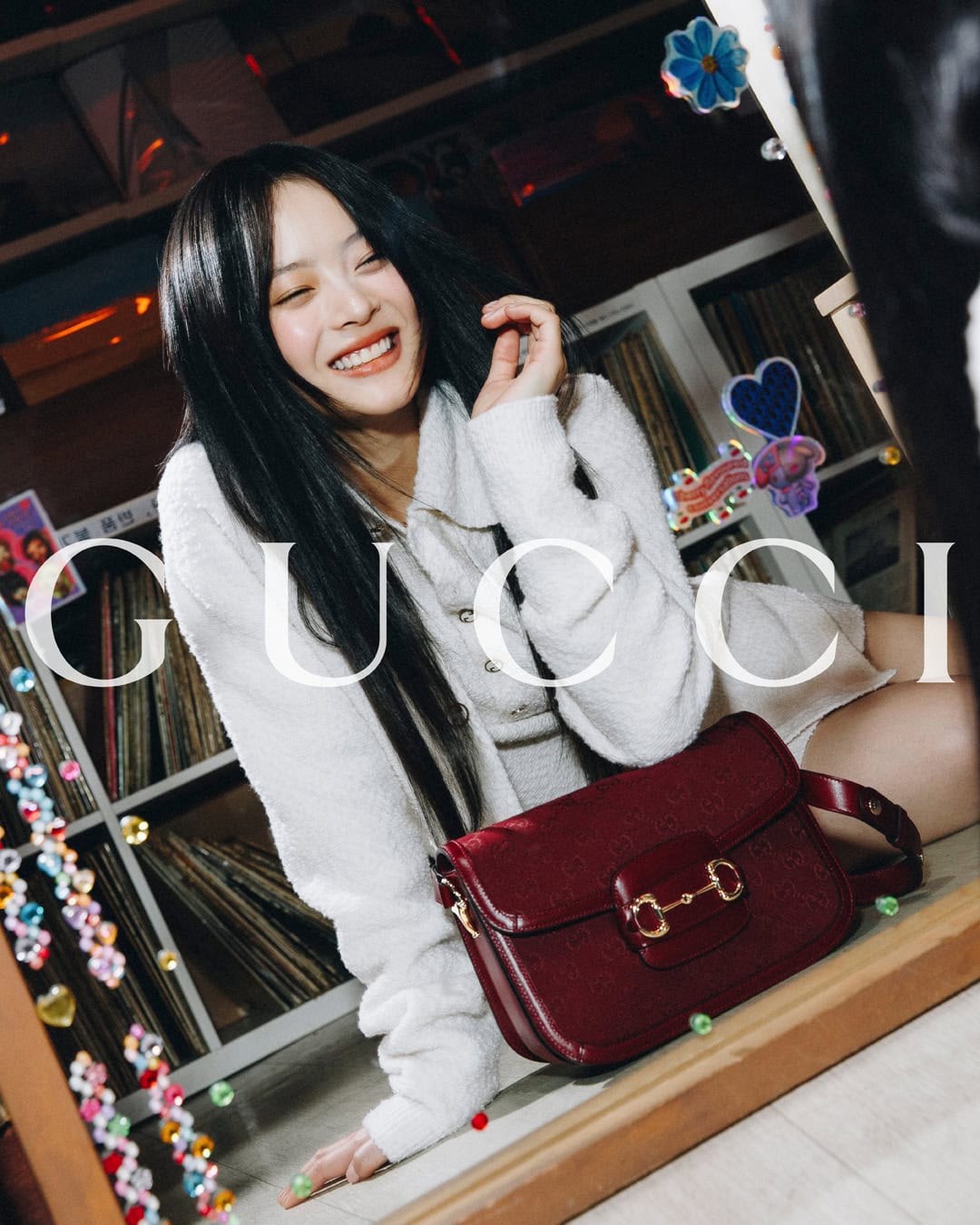 Smiling Asian woman with long black hair wearing white jacket and burgundy Gucci bag in front of record collection.
