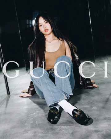 Young woman poses sitting indoors wearing a brown jacket, beige top, blue jeans, and black Gucci loafers with gold hardware.