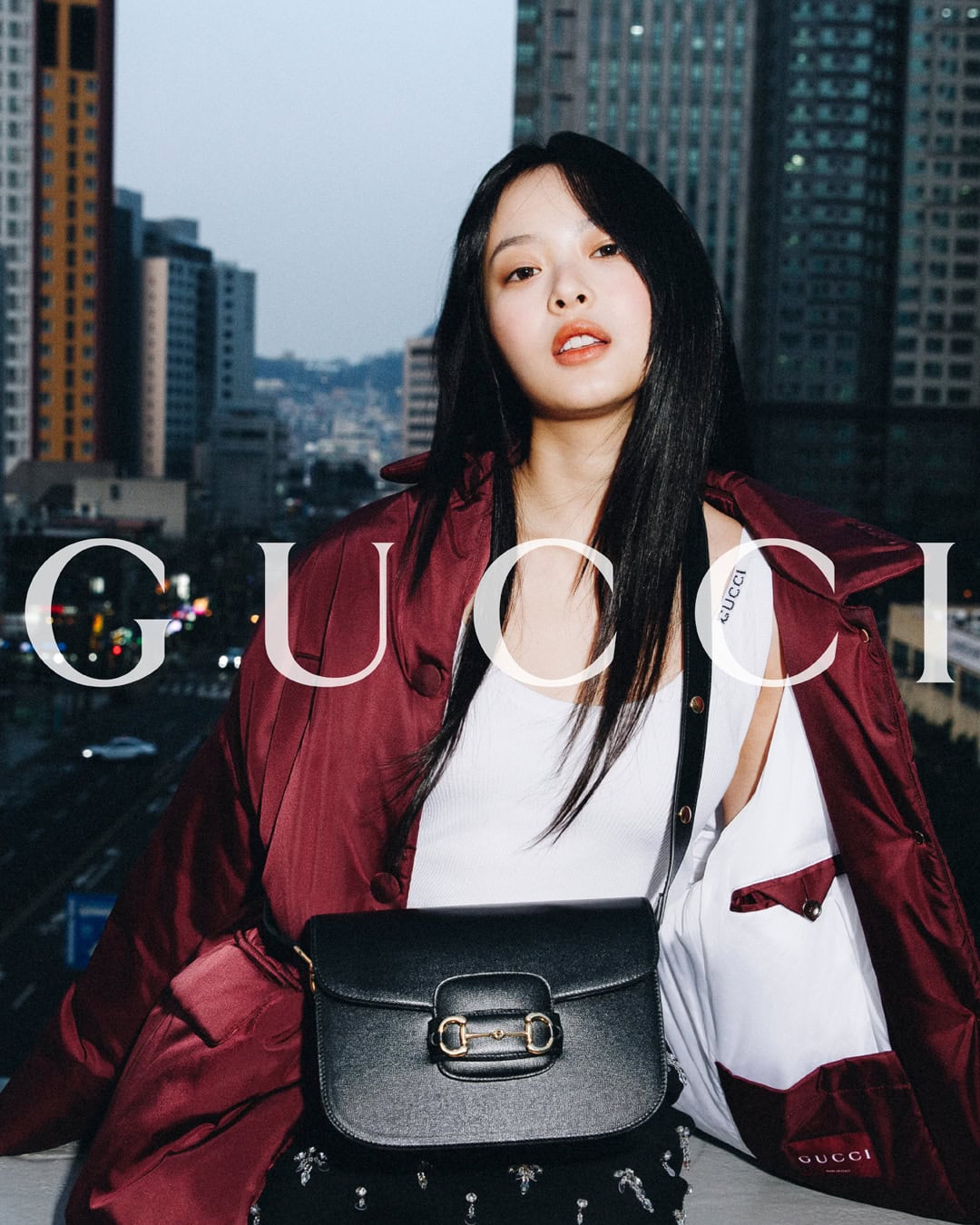 Hanni for Gucci Horsebit Summer 2024 campaign wearing a burgundy jacket and white top with a black handbag against an urban skyline.