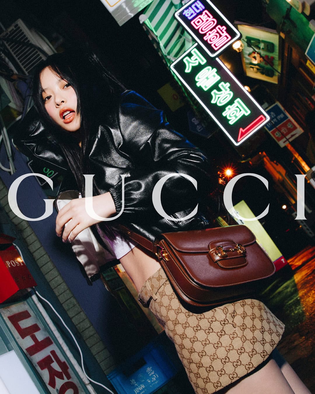 Woman in Gucci monogram dress and leather jacket with a brown handbag, posing in front of neon signage at night. Fashion editorial.