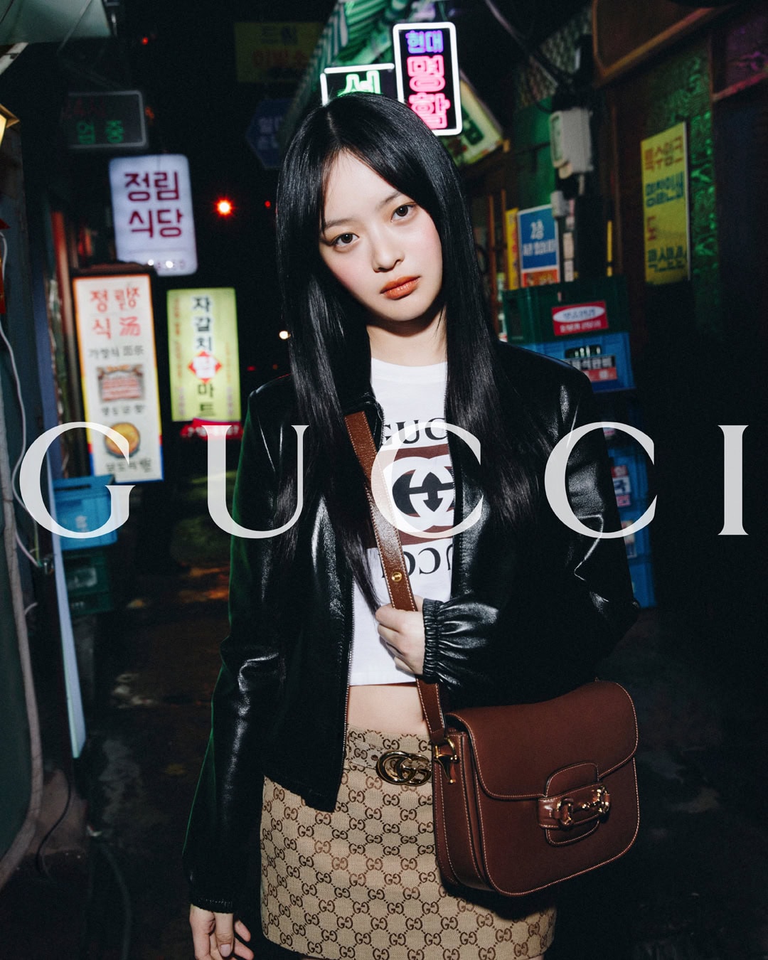 Young woman poses in a black leather jacket and patterned mini-skirt against the backdrop of a city alley with neon signs.
