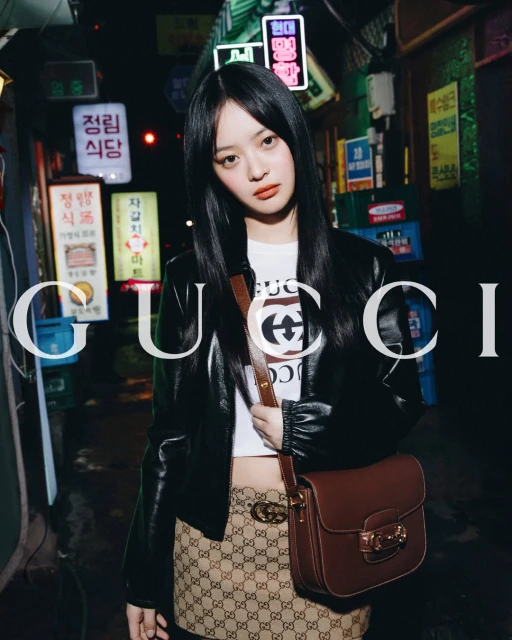 Young woman poses in a black leather jacket and patterned mini-skirt against the backdrop of a city alley with neon signs.
