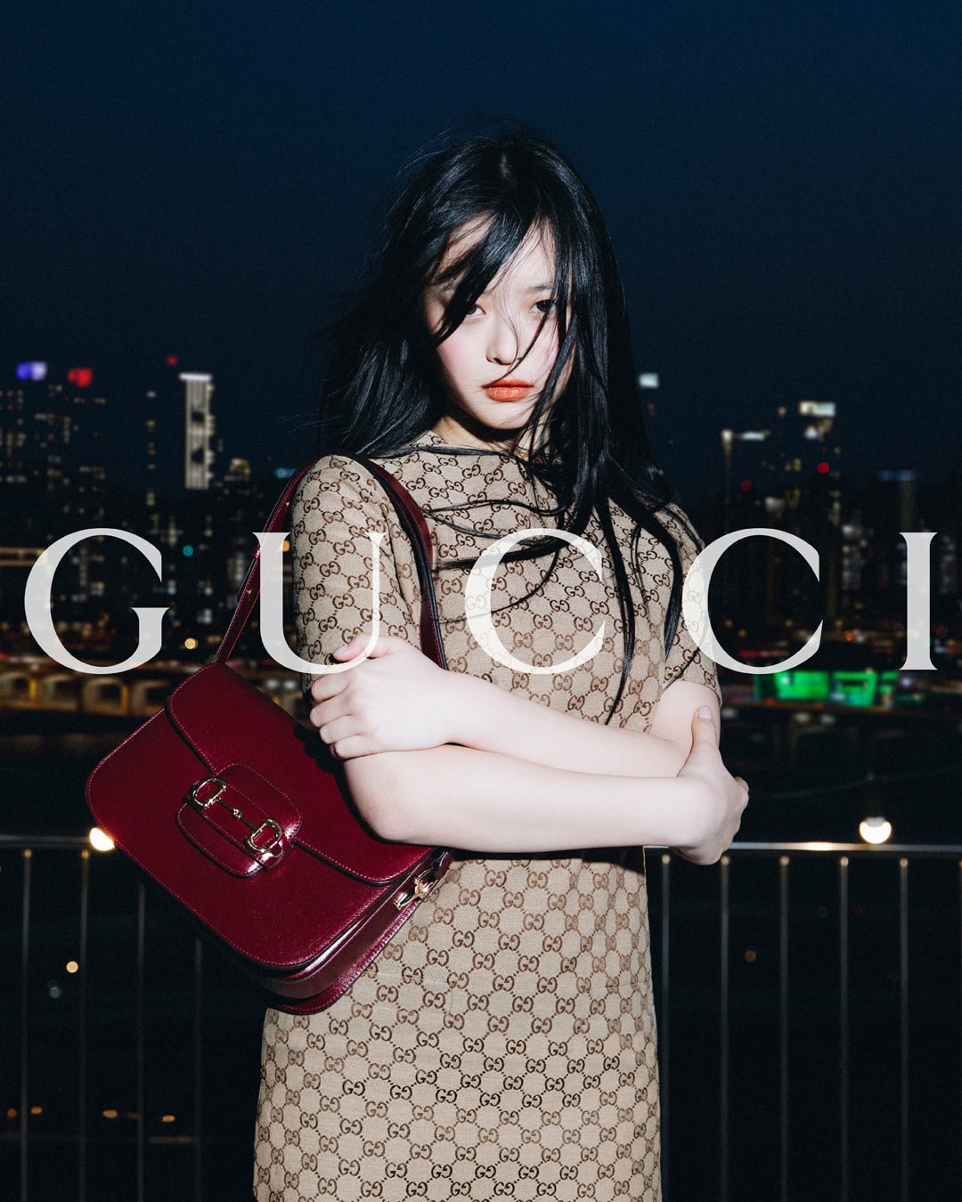 Adult female with a Gucci bag, dress, and long black hair, posing against an illuminated cityscape at night in a medium shot.
