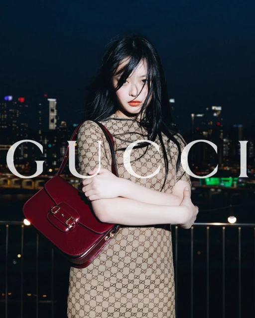 Adult female with a Gucci bag, dress, and long black hair, posing against an illuminated cityscape at night in a medium shot.