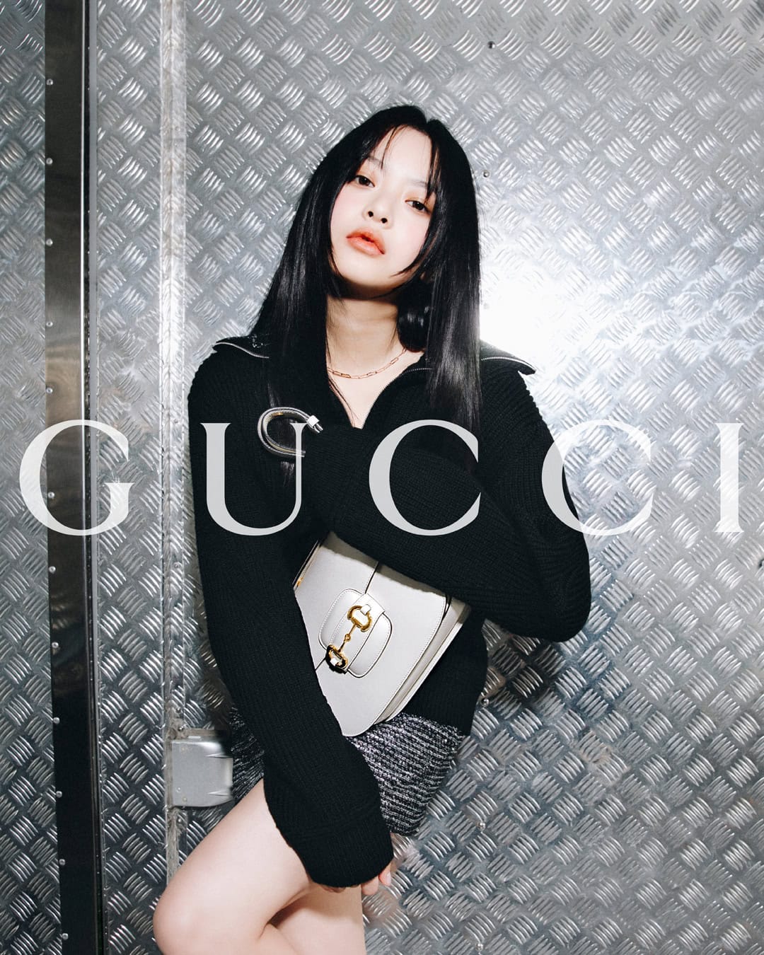 A woman poses with a purse in front of a metal wall, wearing a black sweater and grey skirt. Large Gucci logo is laid over the image.