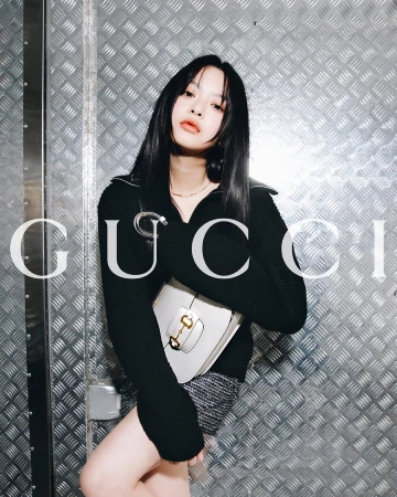 A woman poses with a purse in front of a metal wall, wearing a black sweater and grey skirt. Large Gucci logo is laid over the image.