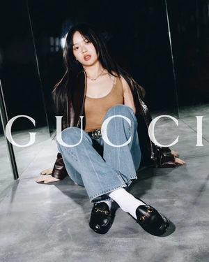 Hanni seated on a dark concrete floor wearing light wash denim jeans, brown tank top, and black loafers with Gucci horsebit hardware
