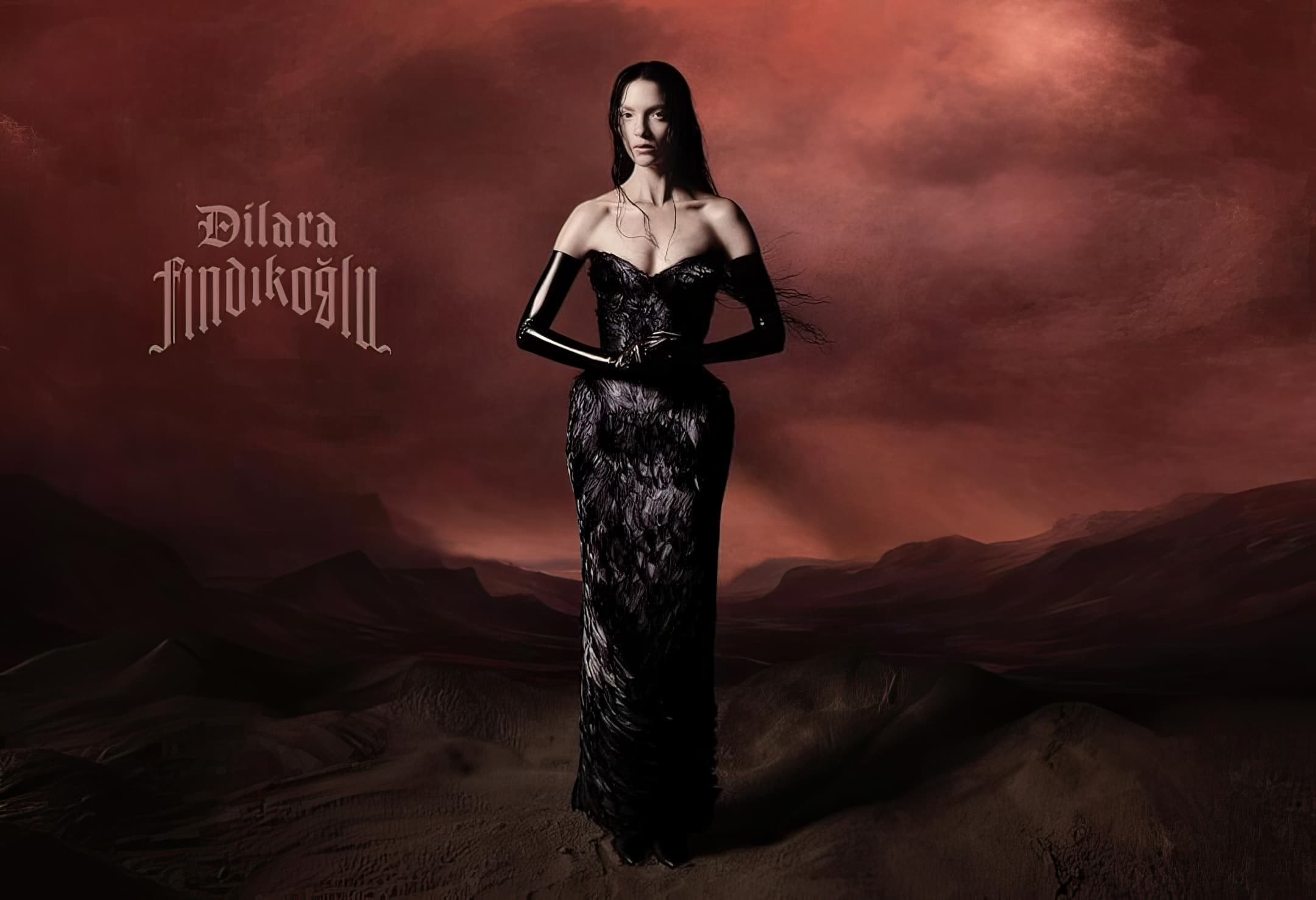 An adult female poses in a black strapless feather gown in a desert setting with reddish-brown hills and sky for a fashion editorial.