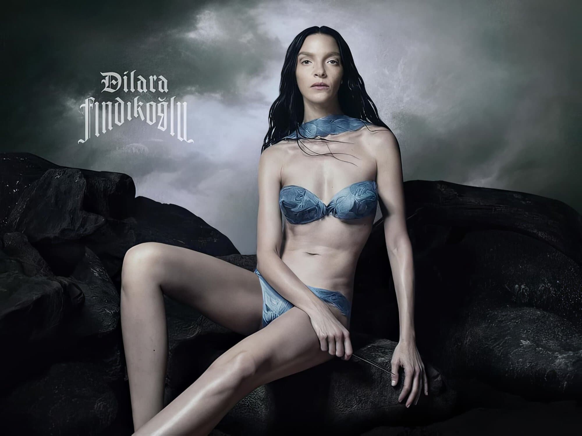 A slender woman with long dark hair poses in a blue bikini, seated on rocks with a cloudy sky behind her in an editorial fashion shot.