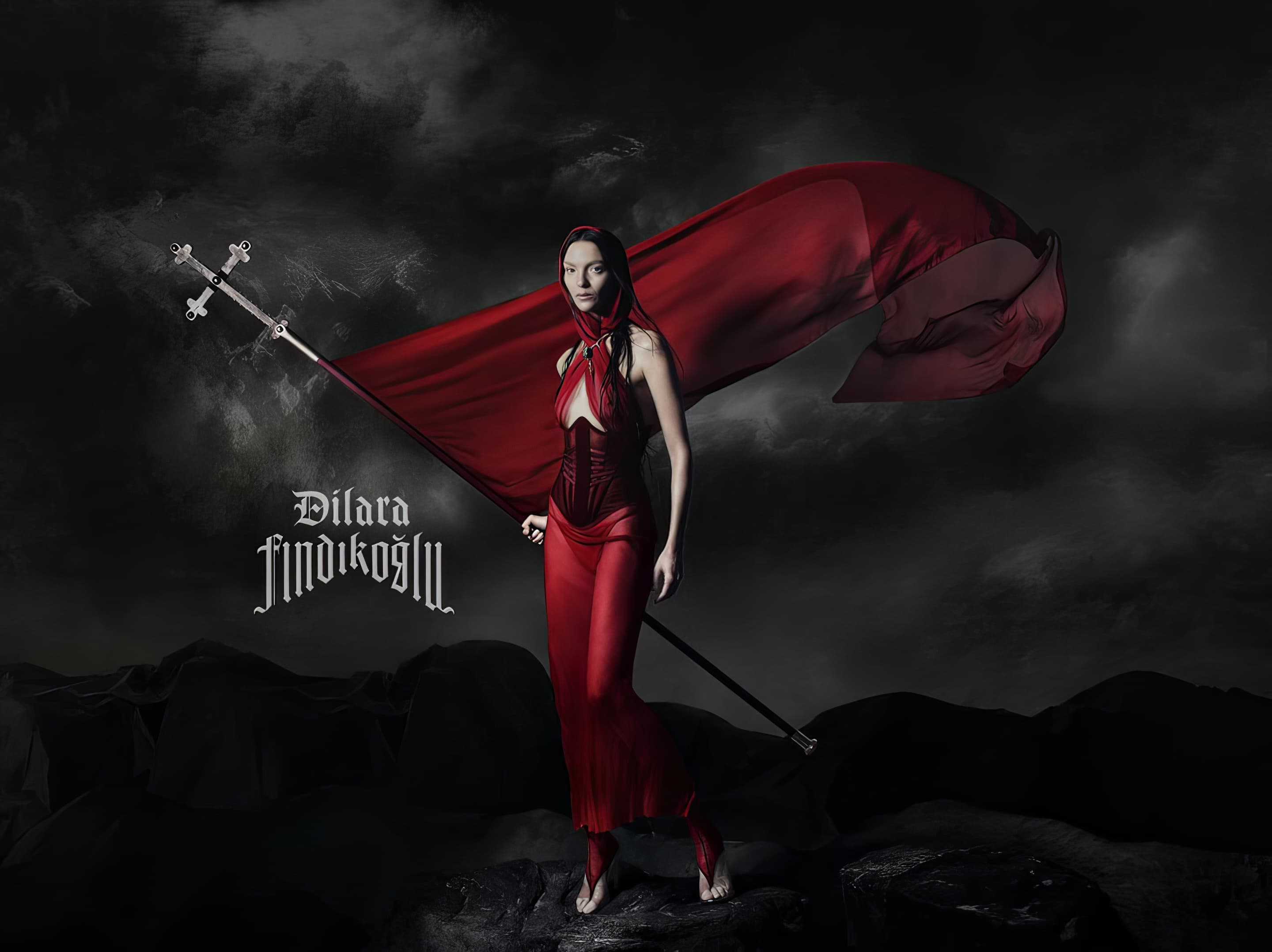 An adult female stands in a draped red gown holding a cross-topped flag against a dark landscape with cloudy skies in a fashion editorial.