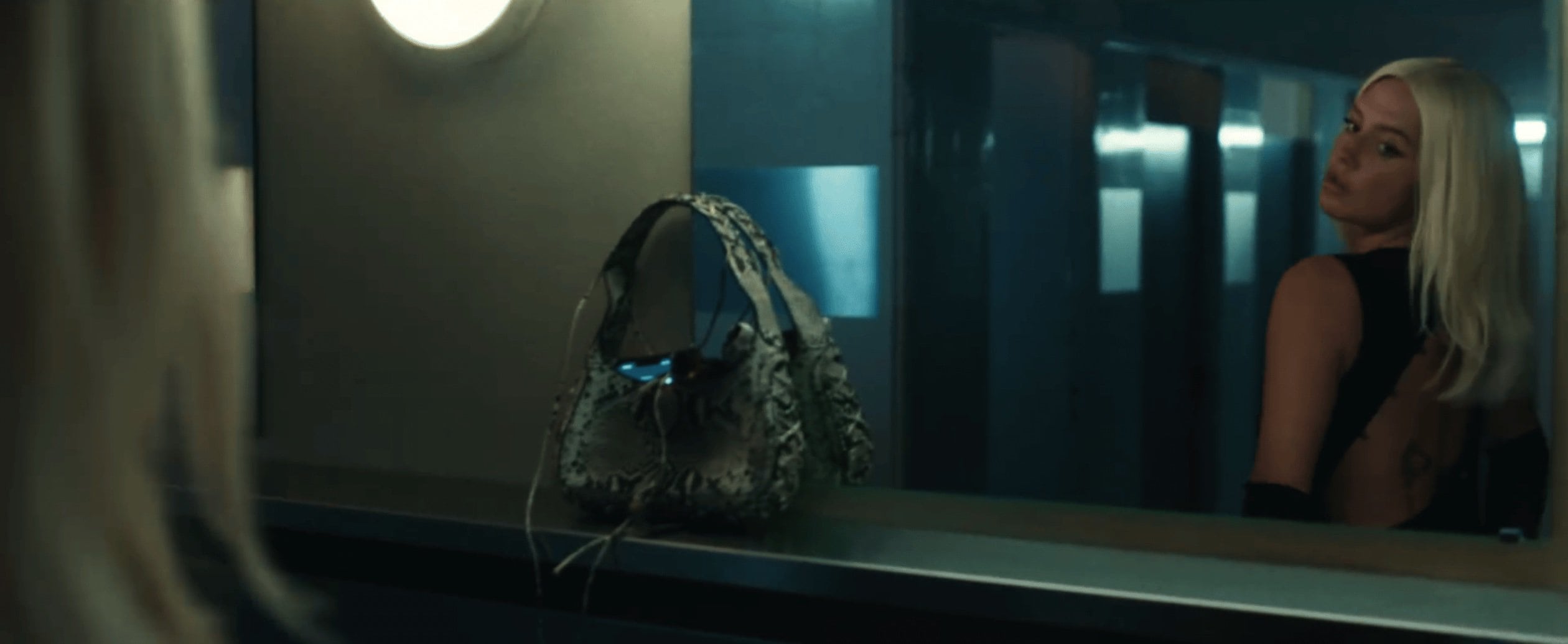 Blonde woman with a black top looks over her shoulder beside an animal print bag in a dimly lit setting with a metallic background.