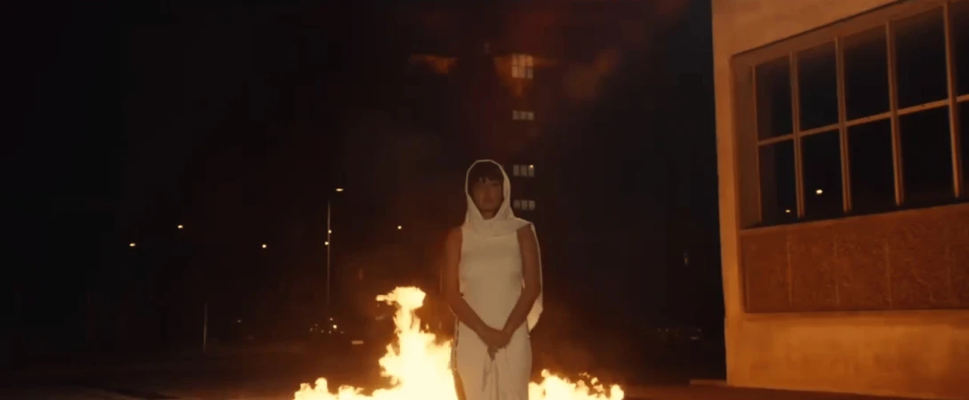 Person in a hooded garment stands before a fire at night, with a building facade and dim city lights visible in the background.