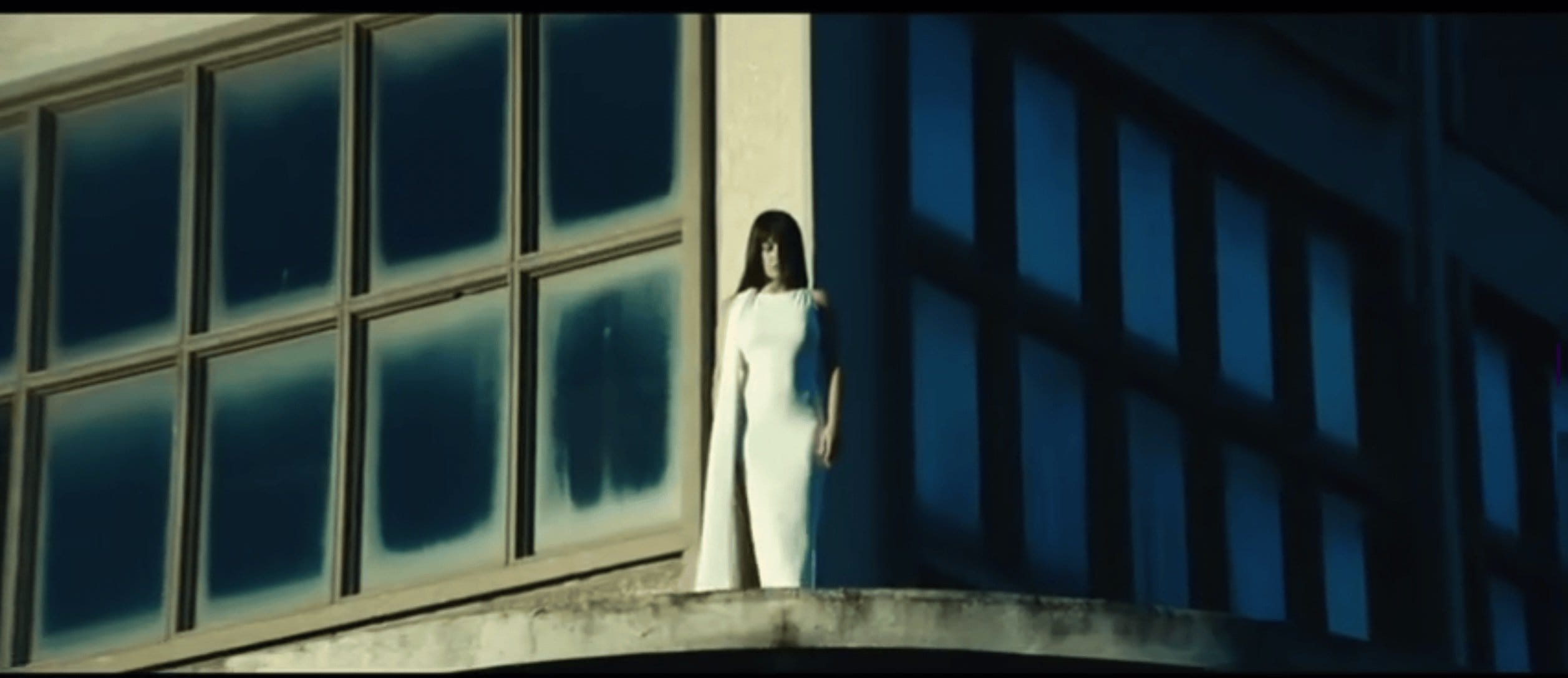 Woman in white dress stands on a balcony next to a large window with a blue tinted reflection in an outdoor setting.