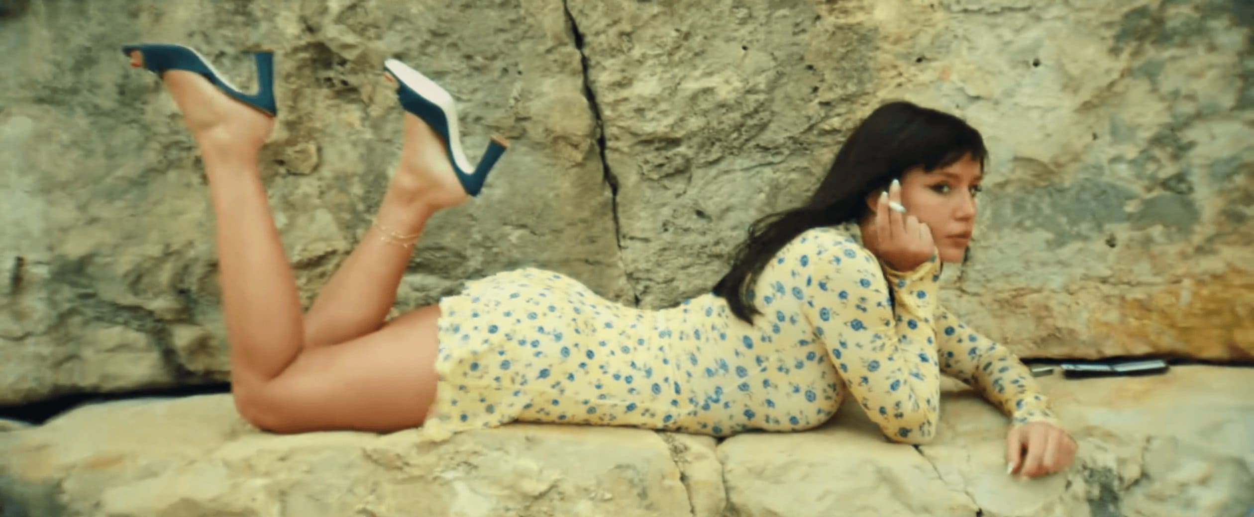 Woman in floral dress and heels poses on rock surface outdoors. Sunlight highlights her features and the natural textures around her.