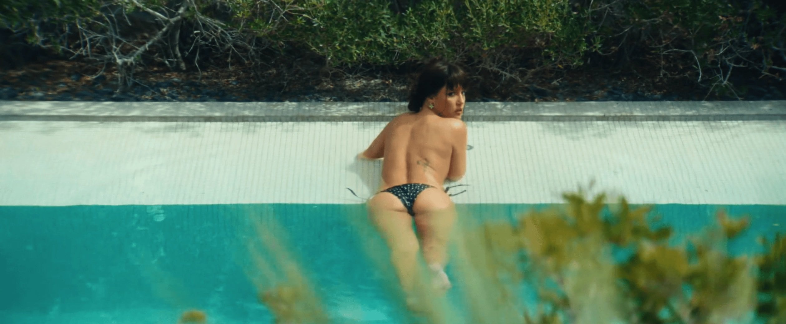Rear view of adult woman in polka-dot bikini lounging at the edge of a bright blue swimming pool surrounded by lush green foliage.