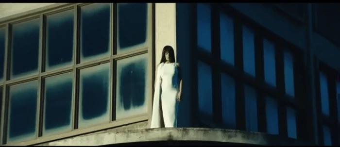 Model wearing a white dress standing on a concrete rooftop against a building exterior with large gridded windows.