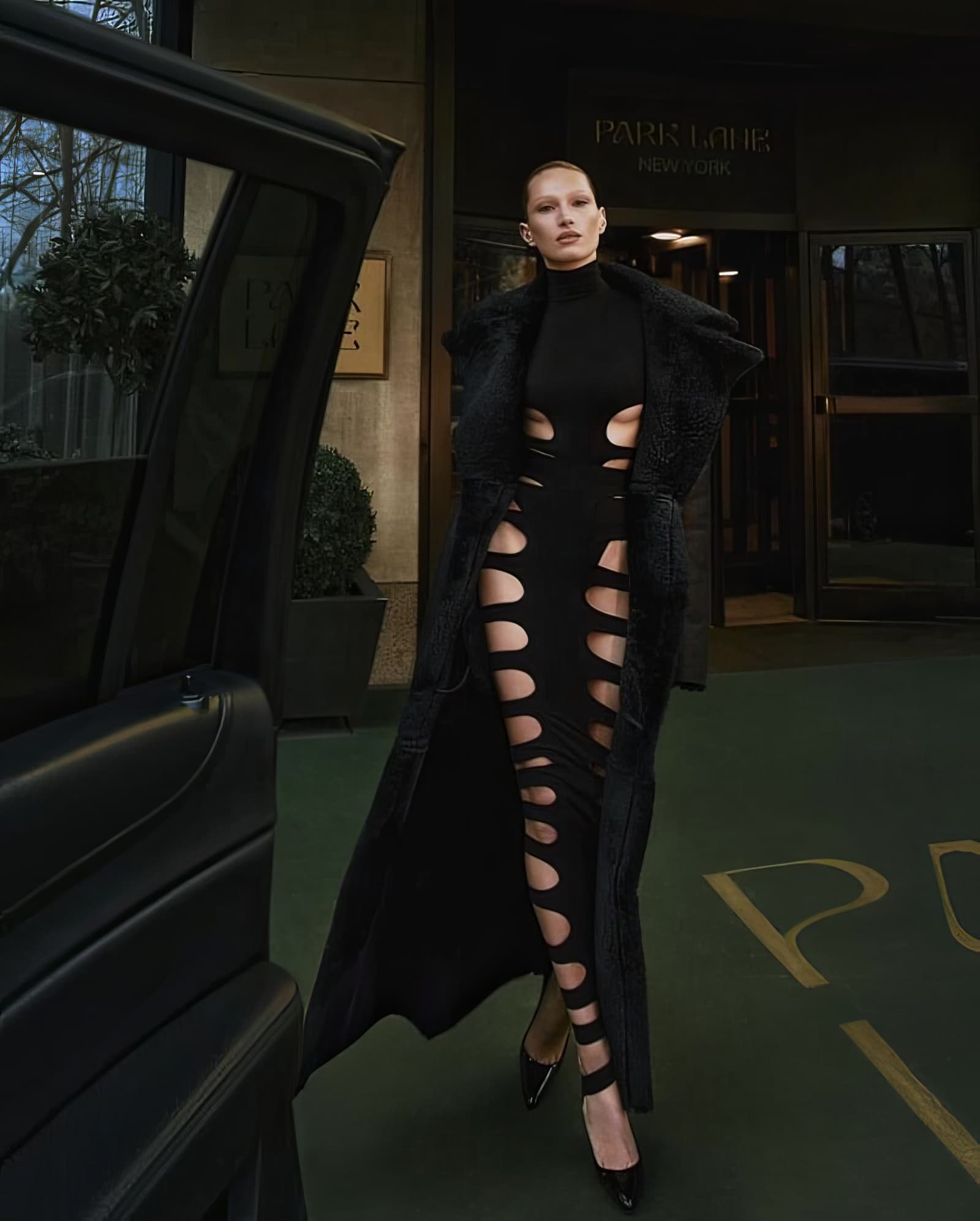 Elegant woman in a black cut-out dress and long coat stands outside a hotel, near a black car, creating a fashion-forward editorial scene.