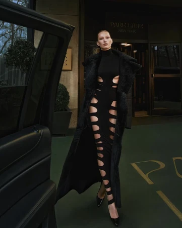 Elegant woman in a black cut-out dress and long coat stands outside a hotel, near a black car, creating a fashion-forward editorial scene.