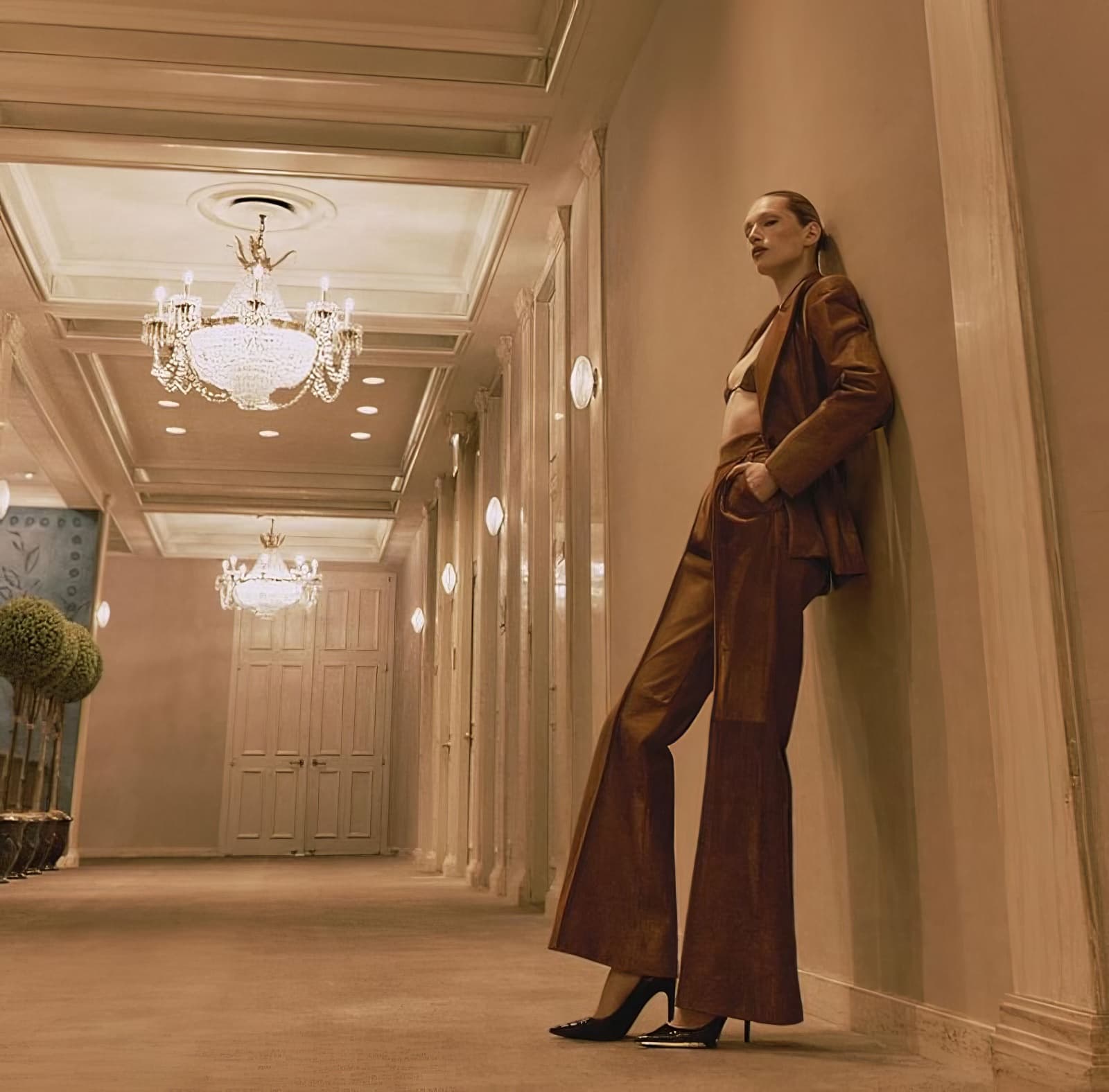 Elegant woman in brown suit posing in a hallway with chandeliers, leaning against the wall in a fashionable pose.