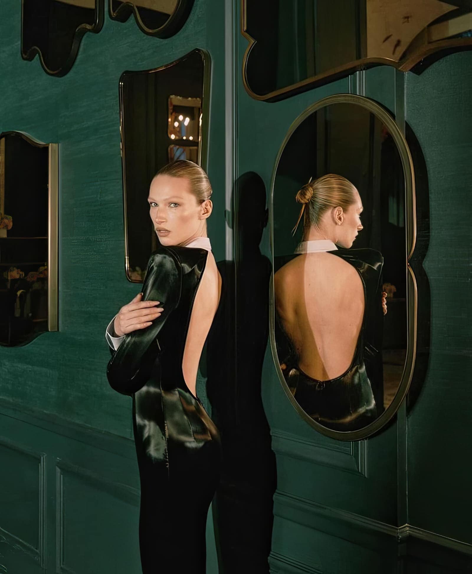 Blonde woman in a black, backless dress posing in front of a green wall with decorative mirrors in an indoor environment.
