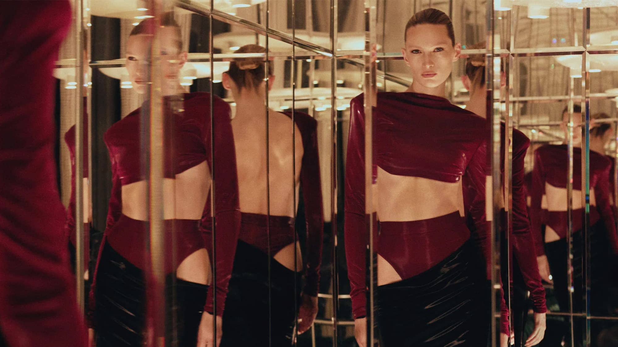 Fashion shot of a slender adult woman in a red top and black skirt posing in a mirror-filled indoor environment with lights.