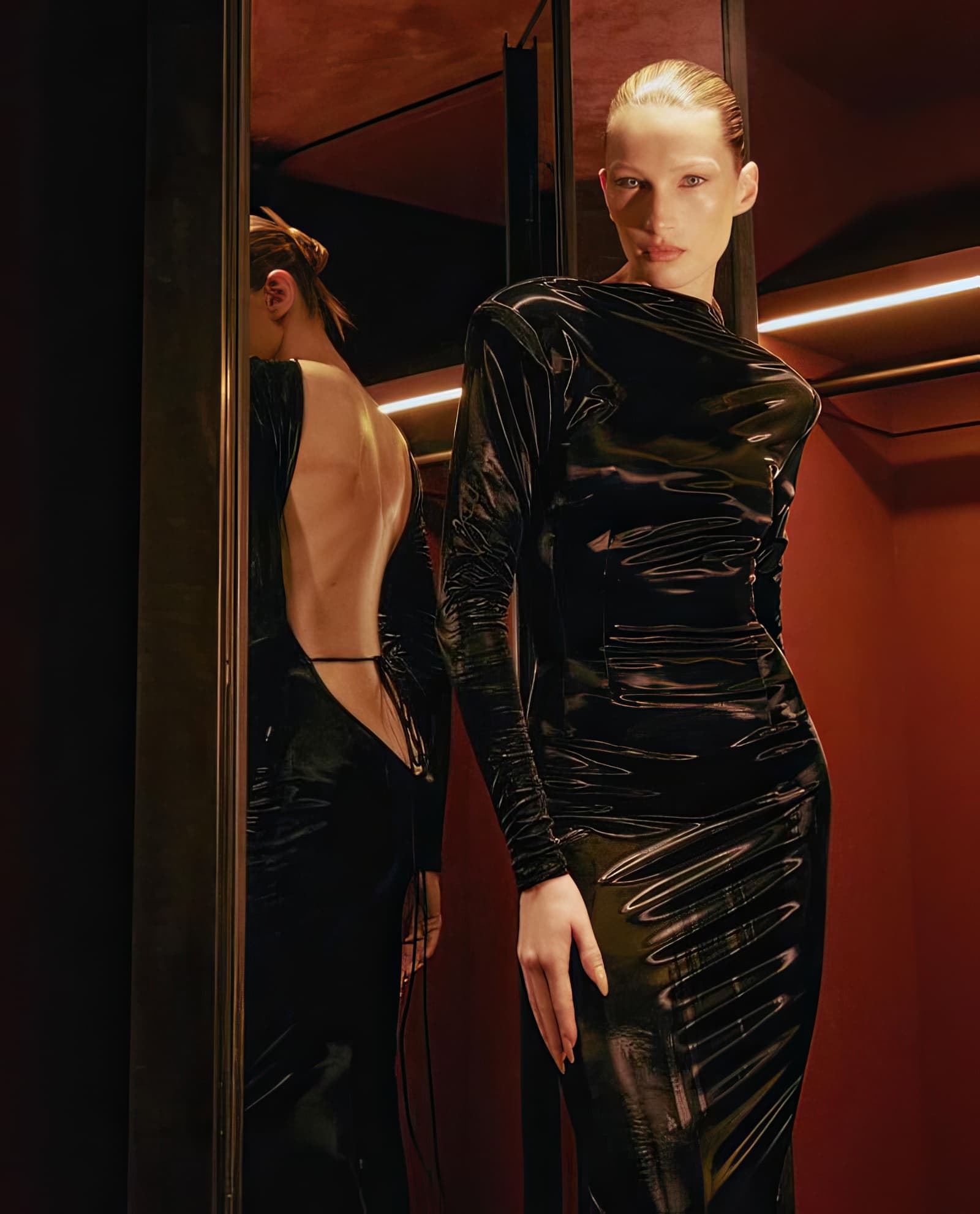 Blonde female model in a black shiny dress poses in front of a mirror in a warm-toned indoor setting, showcasing fashion and style.