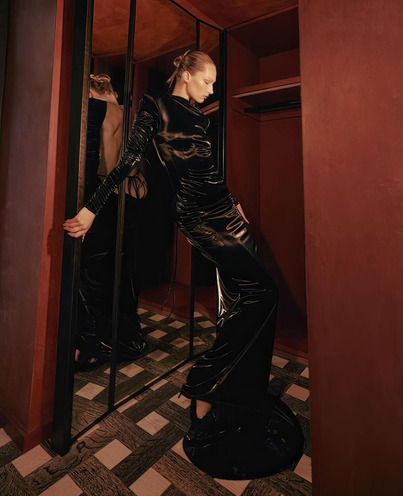 A slender woman in a black, patent-leather-look outfit leans against a reflective column in an interior setting with warm, muted tones.