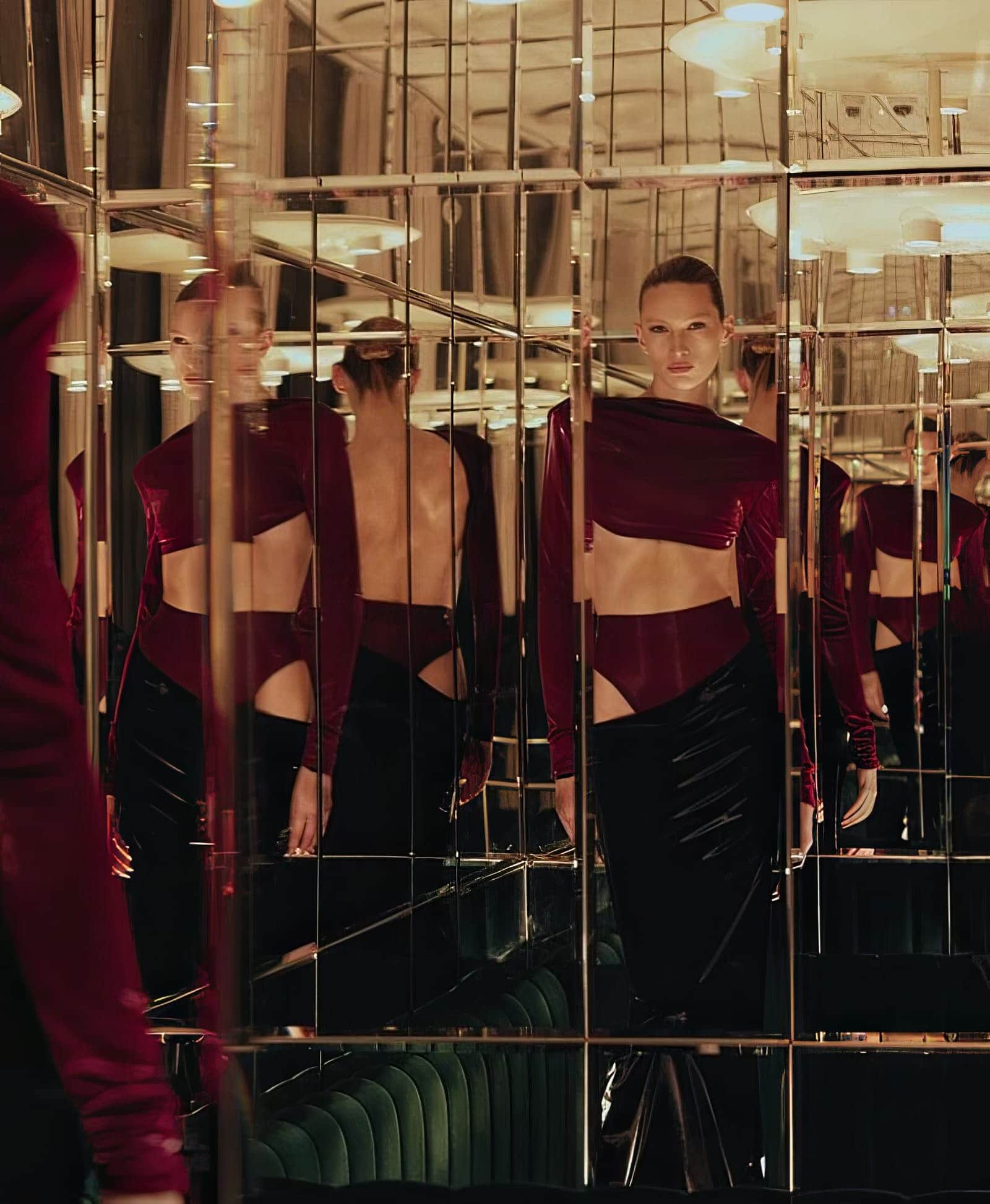 Woman in a modern burgundy top and black skirt poses in a mirror room with a green vintage sofa and modern lights reflecting.
