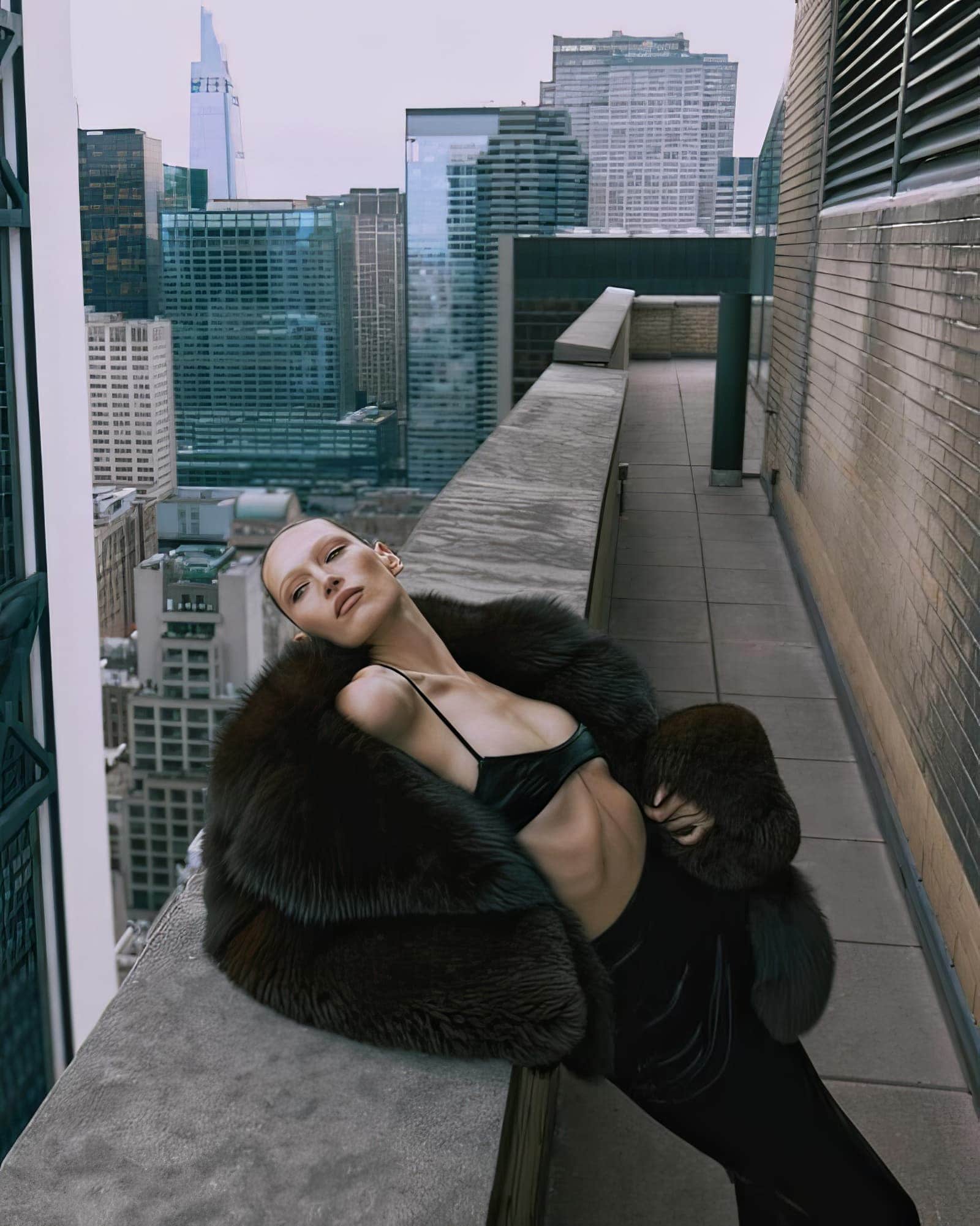 Fashion shot of a slender adult woman posing on a rooftop in a fur coat and velvet clothing with a cityscape in the background.