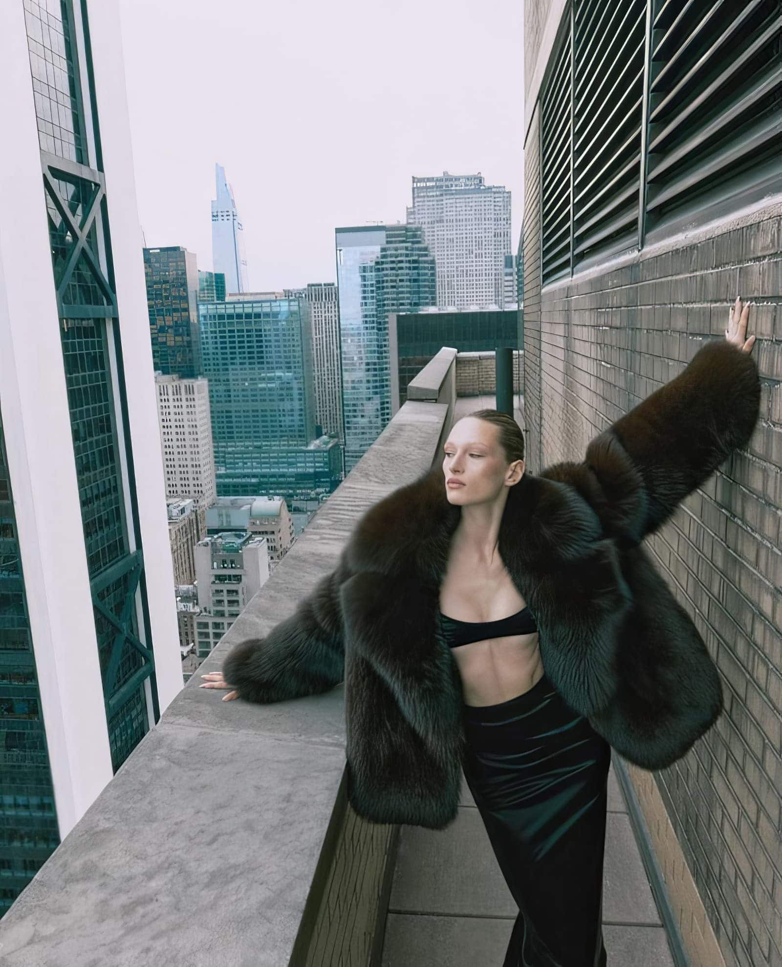 Model standing on a high-rise rooftop overlooking a city skyline, wearing a dark fur coat and black outfit.