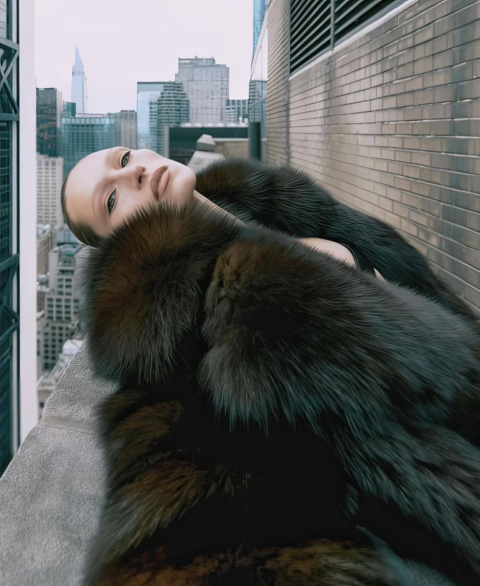 Fashion portrait of a fair-skinned adult woman in a fur coat lying on a rooftop with a city skyline in the background.