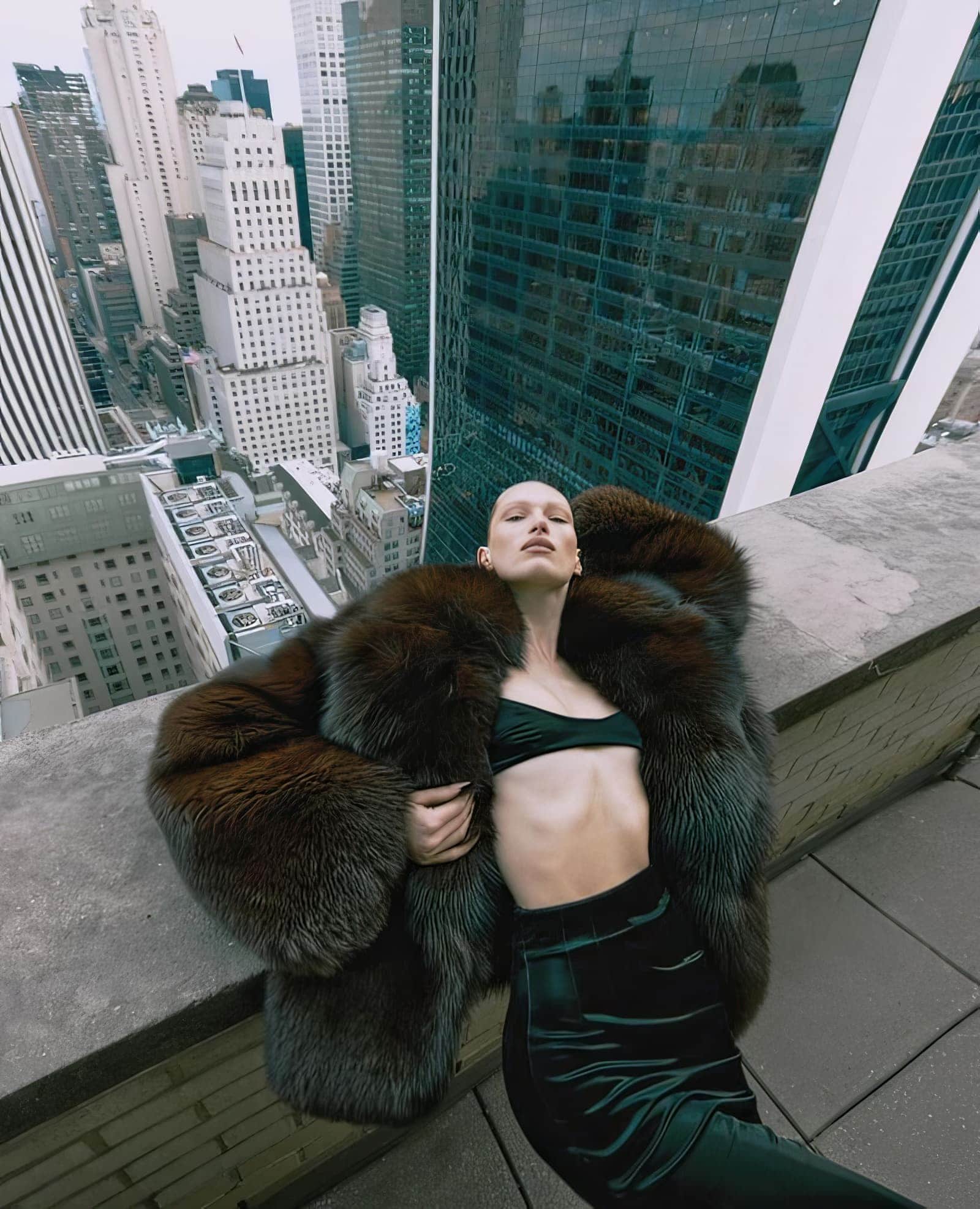 Fashion shot of a woman in a fur coat and velvet skirt lying on a rooftop with a city skyline in the background on an overcast day.