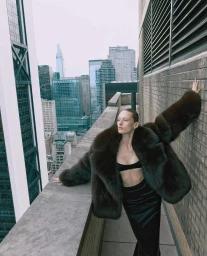Model standing on a high-rise rooftop overlooking a city skyline, wearing a dark fur coat and black outfit.