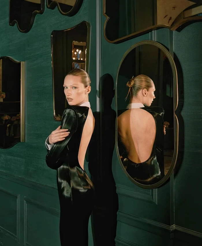 Model Liz Kennedy wearing a backless black dress posing against a green wall with mirrors for LaQuan Smith Fall 2024 Ad Campaign.