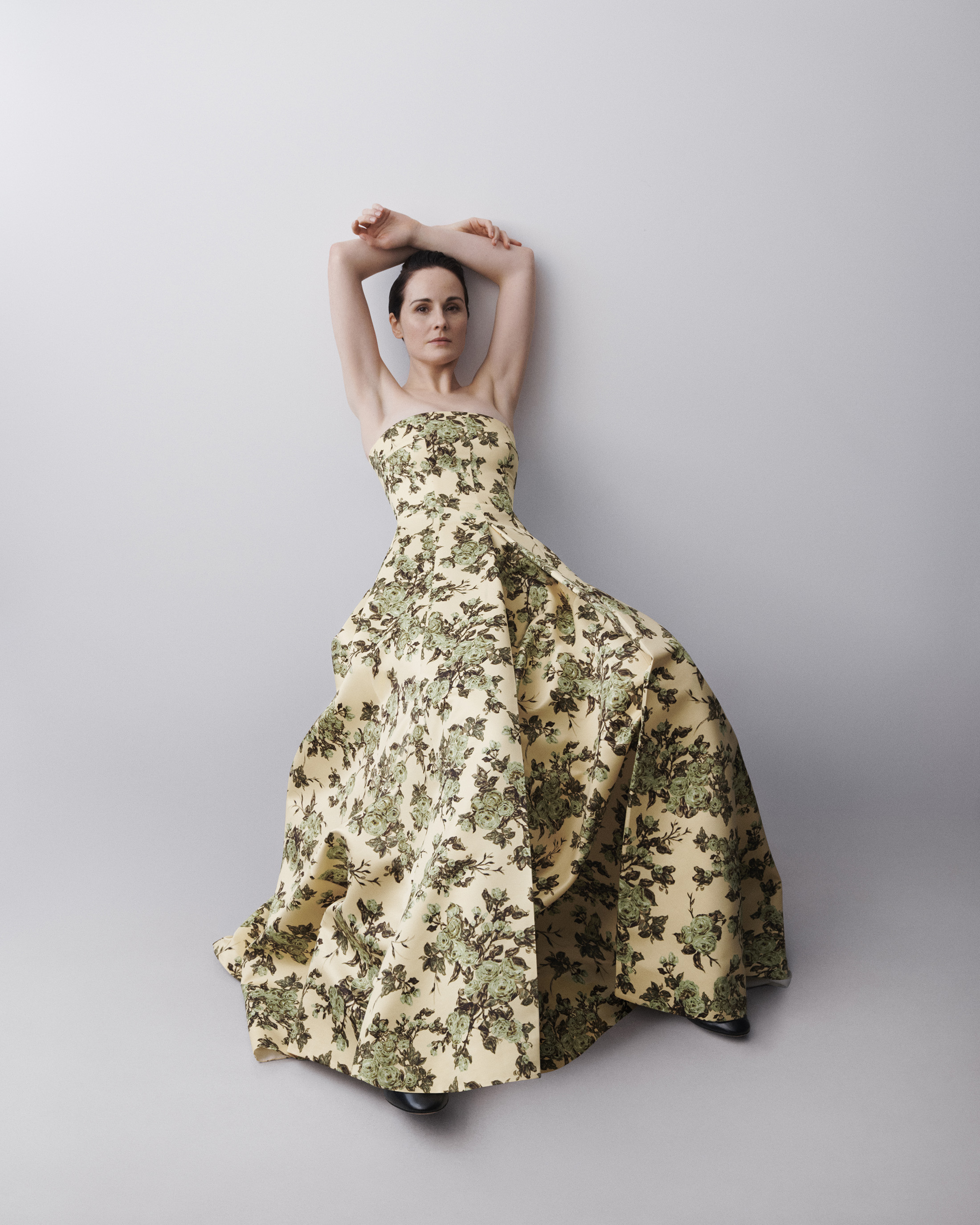 Woman poses in floral strapless gown against a light gray wall, arms raised above her head, looking at the camera in a fashion shot.
