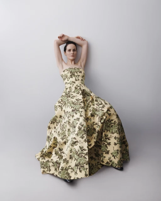 Woman poses in floral strapless gown against a light gray wall, arms raised above her head, looking at the camera in a fashion shot.