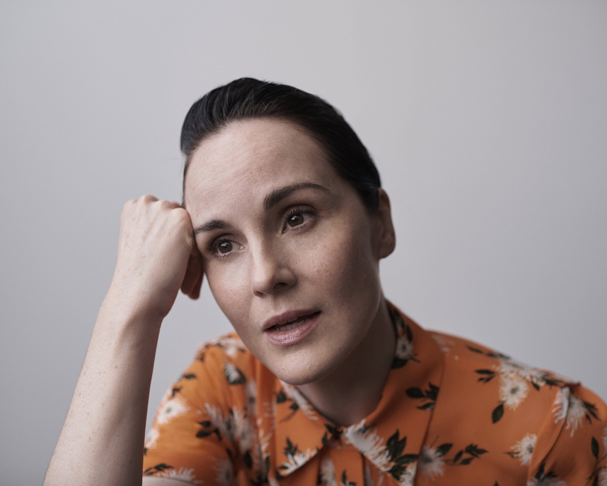 Portrait of an adult woman with dark hair, wearing an orange floral blouse, resting her head on her hand in a photo studio setting.