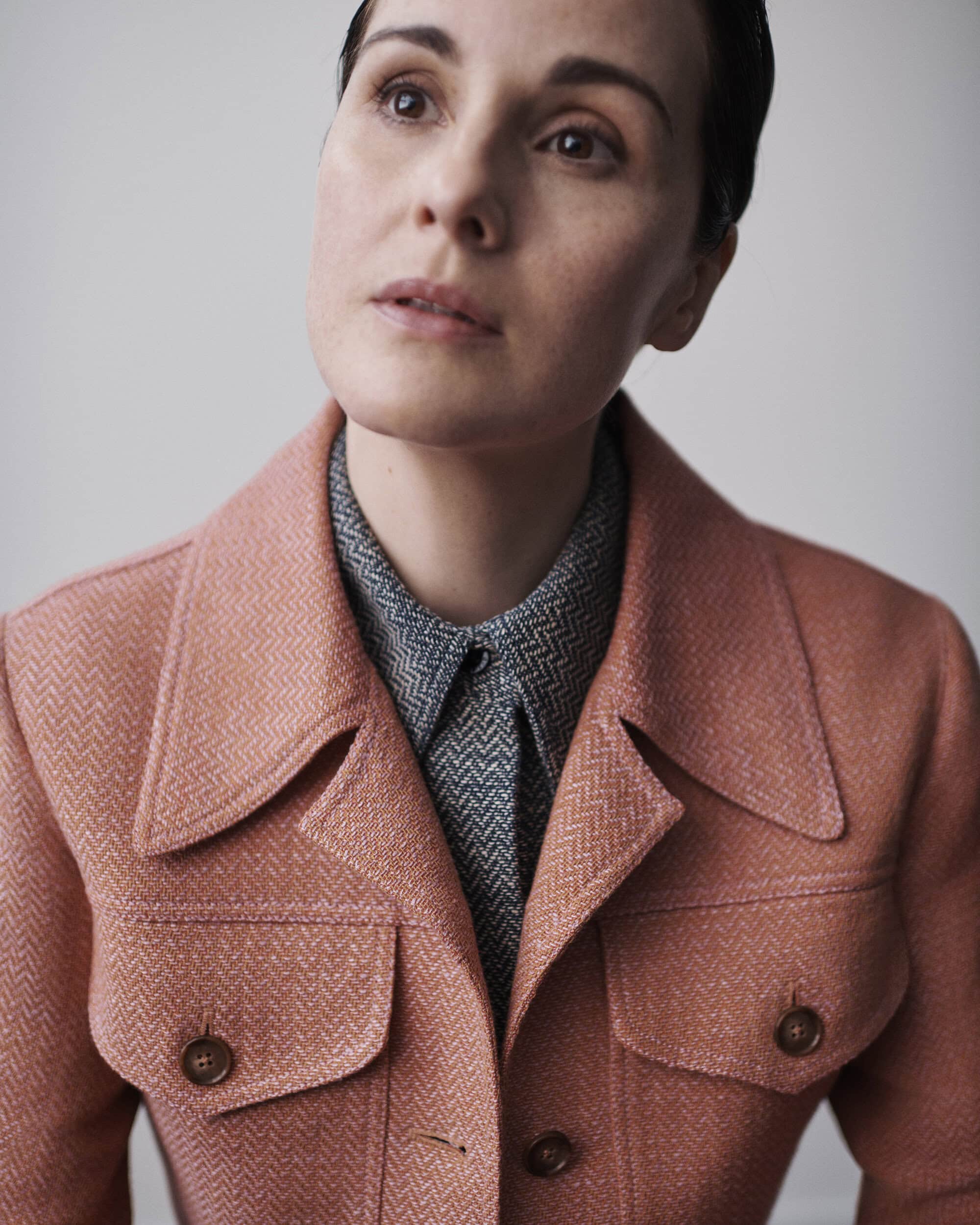 Portrait of a woman with brown hair wearing a pink jacket over a patterned grey shirt, looking up against a neutral background.