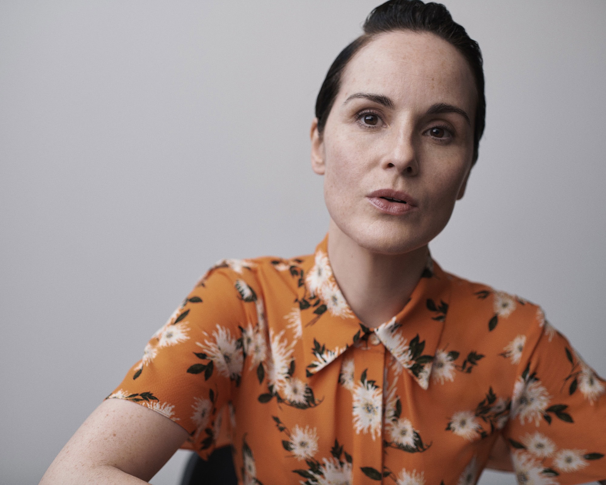 Portrait of a dark-haired woman wearing an orange, floral print blouse, looking directly at the camera with a neutral expression.