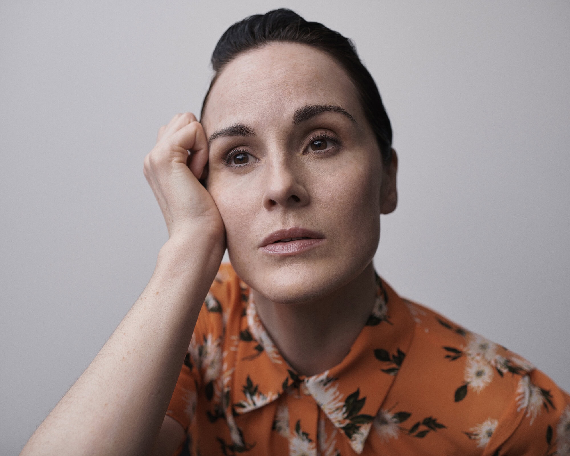 Portrait of an adult woman with dark hair in an orange floral shirt, resting her head on her hand, looking upwards in soft lighting.