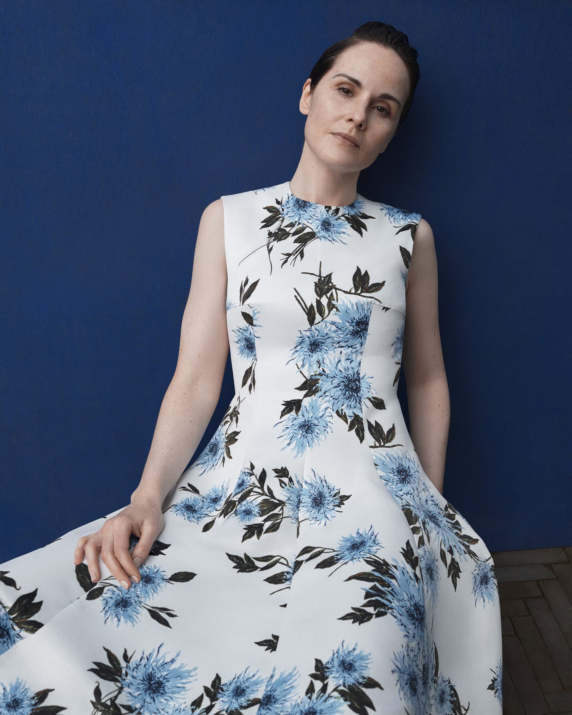 Elegant woman in a sleeveless white dress with a blue floral pattern poses against a dark blue wall in a softly lit indoor setting.