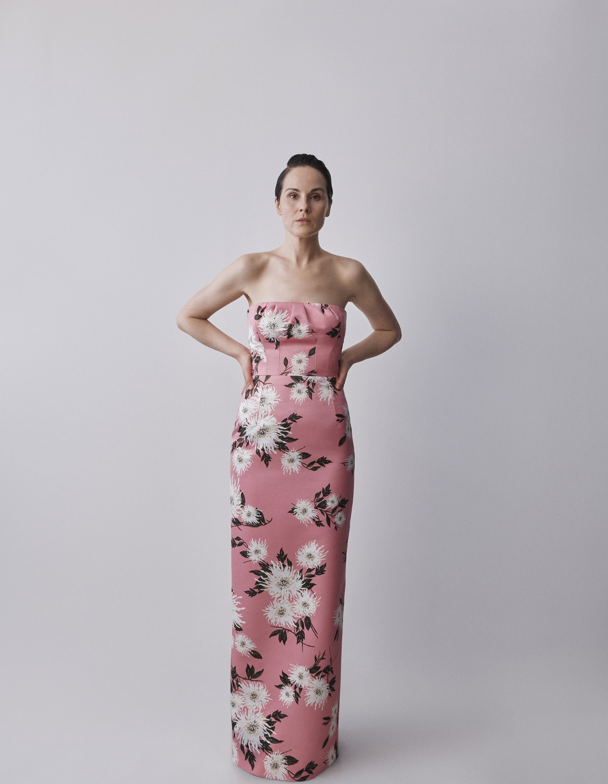 Adult female posing in a long, strapless pink gown with white floral pattern against a plain background in a studio setting.